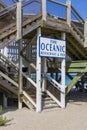 A white and blue sign that reads Ã¢â¬ÅThe Oceanic Restaurant and PierÃ¢â¬Â on a brown wooden pier with people, brown sand